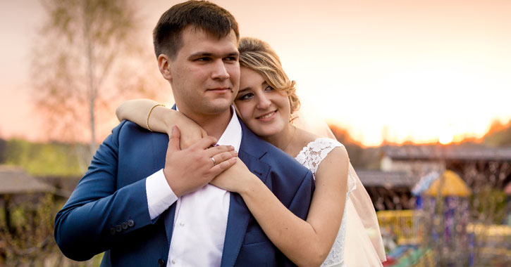 a bride hugging the groom from behind, sunset in the background